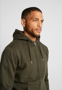 Olivgrüne Zip-Hoodie mit weichem Stoff, Kordelzugkapuze, silberner Hardware und vorderen Taschen. Schlichtes Design mit verstärkten Nähten.
