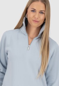 Felpa con zip di colore azzurro chiaro, con colletto alto, texture morbida e design minimalista. Presenta un dettaglio della cerniera in argento e maniche lunghe.