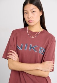 Camiseta corta de manga de color burdeos de Nike con una gran impresión de logo gris. El modelo tiene el cabello largo y negro y usa collares en capas. Textura de tela suave.