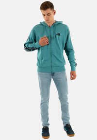 Giovane uomo che sistema la felpa con zip color turchese e strisce nere sulle maniche, indossando jeans azzurri e scarpe da ginnastica blu scuro con strisce blu.