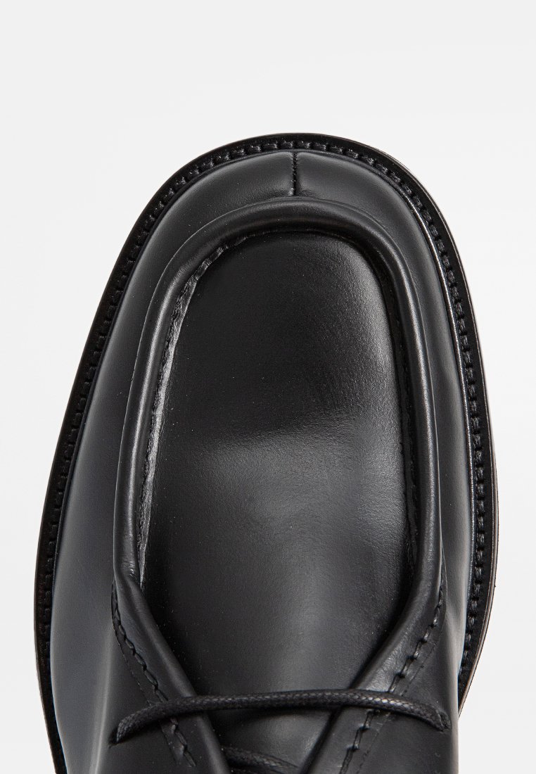 Zapato de cuero negro con una punta redondeada y lisa, detalles cosidos y cordones negros. El borde de la suela está acabado con una costura en contraste.