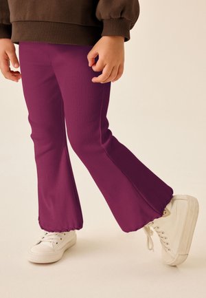 Kind trägt lila ausgestellte Hose, weiße Sneakers und ein braunes langärmeliges Oberteil, steht mit einem leicht angehobenen Fuß.