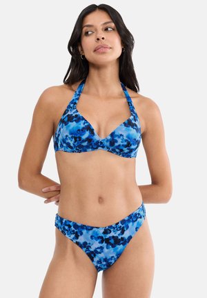Kvinde iført blå og sort floral halter-bikinitop og matchende højt udskåret bikinitrusser, stående med armene bag ryggen.