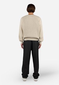 Man standing facing away wearing a cream ribbed sweater, black wide-leg pants, and light-colored shoes on a white background.
