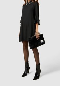 Schwarzes Kleid mit ausladenden Ärmeln und spitzem Kragen, kombiniert mit einer flauschigen schwarzen Handtasche und glänzenden Knöchelstiefeln. Strukturiertes, modernes Design.