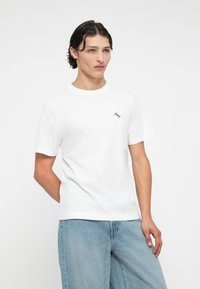 White cotton t-shirt featuring a round neck, short sleeves, and a small embroidered logo on the left chest. Paired with light blue denim.