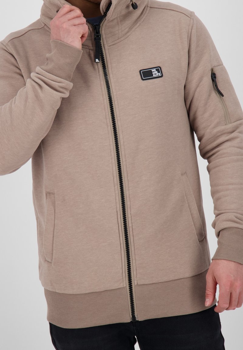 Uomo che indossa un hoodie beige con zip e un patch "WE ARE ALIFE", mano vicino al collo, e pantaloni neri su sfondo neutro.