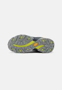 Semelle Vibram avec un motif gris robuste orné d'accents jaunes et de crampons profonds. Partie supérieure noire et violette visible au talon.