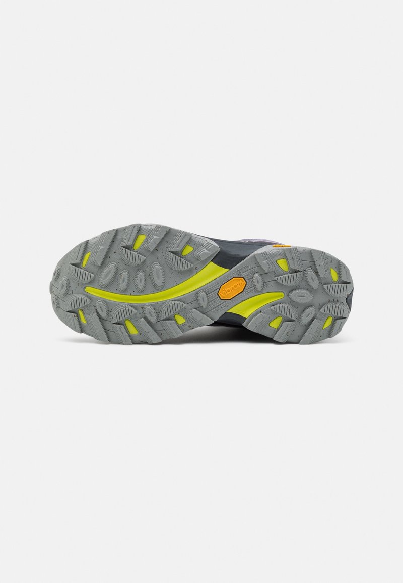 Semelle Vibram avec un motif gris robuste orné d'accents jaunes et de crampons profonds. Partie supérieure noire et violette visible au talon.