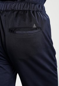 Pantaloni jogging blu navy con vita elasticizzata, dotati di una piccola tasca laterale con un logo triangolare e una texture di tessuto liscia e leggera.
