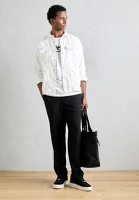 Giacca in denim bianca sopra una t-shirt bianca con grafica nera, pantaloni neri e sneakers nere con suola bianca, mentre tiene una borsa a tracolla nera.