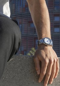 Reloj de acero inoxidable plateado con una carátula blanca y acentos azules, eslabones rectangulares y un acabado pulido. Lleva puesto en una muñeca descansando sobre un borde de concreto.