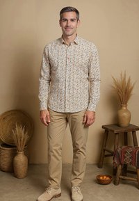 Lichtkleurig overhemd met bloemenpatroon in oranje en groen, gedragen met beige broek en lichte schoenen, tegen een neutrale achtergrond.