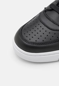 Svart lädersneaker med perforerade detaljer, kontrasterande vit gummisula och texturerad sömnad längs kanterna.