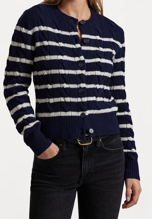 Vrouw draagt een marineblauw-wit gestreepte kabelgebreide vest met knopen en geribbelde manchetten, gecombineerd met zwarte jeans en een zwarte riem.