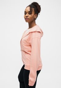 Νεαρή γυναίκα με σγουρά μαλλιά φοράει ροζ φούτερ με φερμουάρ και μαύρο παντελόνι, στάθηκε πλάγια μπροστά σε ένα απλό λευκό φόντο.