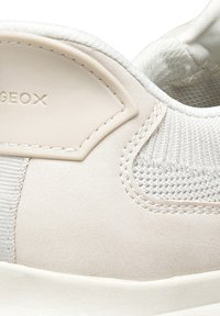 Detailní záběr béžové tenisky s síťovinou, vyšitou výšivkou a gumovou záplatou s vyraženým názvem značky "GEOX" na boku.