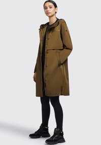 Cappotto impermeabile verde oliva con cappuccio, tasche frontali, chiusura con zip e bottoni a pressione, indossato sopra leggins neri e sneakers nere.
