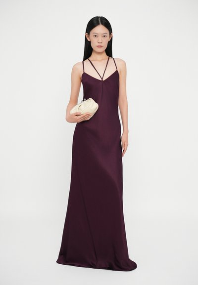 Vestido longo e justo em tecido roxo profundo com alças finas, apresentando um decote em v e uma textura suave e elegante. Modelo segura uma pequena mala bege.