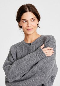 Femeie cu păr închis la culoare, purtând un pulover gri din tricot cu butoane, cu brațele încrucișate și privind spre lateral pe un fundal alb.