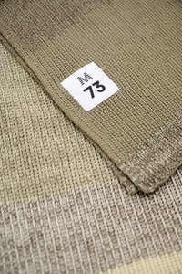 Tissu tricoté côtelé beige avec une étiquette blanche présentant une lettre M rayée et le numéro 73, plié sur un tissu de texture similaire.