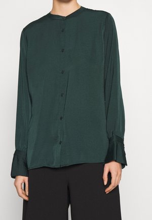 Donkergroene blouse met lange mouwen, knopen en geplooide manchetten, gedragen met zwarte broek, toont bovenlichaam en handen tegen een witte achtergrond.