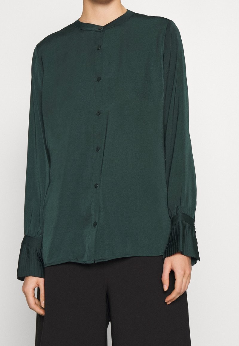 Blouse verte foncée à manches longues avec boutons et poignets plissés portée avec un pantalon noir, montrant le haut du torse et les mains sur fond blanc.