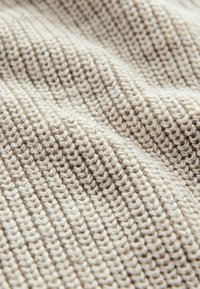 Texturerad stickad tyg i ljus beige och kräm, med ett rytmiskt mönster av växlande stygn och en mysig känsla.