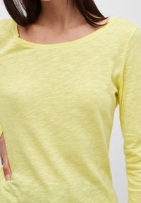 Frau in einem langärmeligen, hellgelben, leicht strukturierten Shirt mit weitem Rundhalsausschnitt, wobei ihre Hand auf ihrer Hüfte ruht.