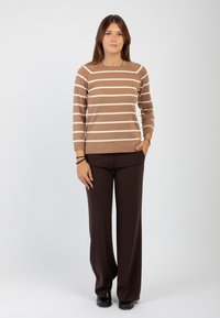 Giovane donna in piedi, che indossa un maglione a righe marroni e bianche, pantaloni larghi marroni scuri e scarpe nere, su uno sfondo chiaro e semplice.