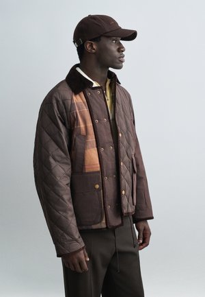 Mand iført brun quiltet jakke med patchwork-detaljer, mørkebrune bukser og matchende brun kasket, vendt mod højre mod en ensfarvet baggrund.