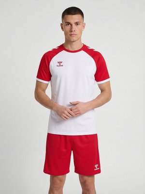 Man staat met gevouwen handen, draagt een wit-rood sportshirt en rode shorts tegen een effen achtergrond.