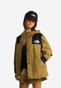 Grøn og sort vandtæt jakke med hætte, forsynet med trykknapper og logo på brystet. Kombineret med beige outfit og sort kasket.