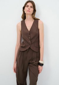 Chaleco sin mangas marrón con cuello en V, botones de un solo pecho y dos bolsillos frontales, combinado con pantalones marrones a juego. Textura suave.