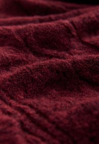 Burgunderroter Wollstoff mit einer weichen, flauschigen Textur, der sanfte Wellen und Falten aufweist. Bekannt für seine kräftige Farbe und warme Ausstrahlung.