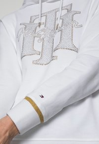 Hoodie bianco con design di strass scintillanti, dettagli dorati sui polsini e un piccolo logo in rosso, bianco e blu sulla manica.