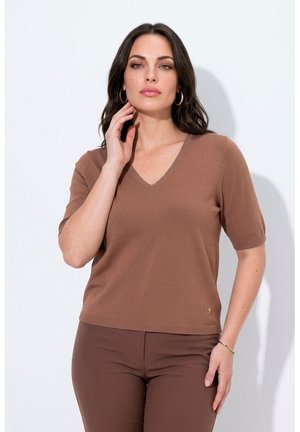Femme aux cheveux foncés portant un haut marron à manches courtes en V et un pantalon assorti, posant avec une main touchant son cou sur fond blanc.