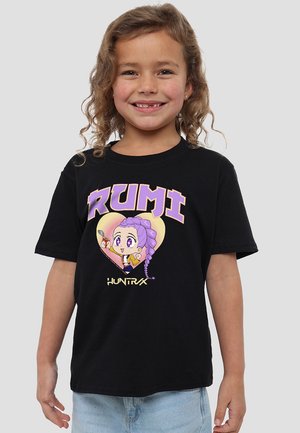 Lächelndes junges Mädchen mit lockigem Haar, das ein schwarzes T-Shirt trägt, auf dem ein lila Cartoon-Charakter sowie die Worte "RUMI" und "HUNTR/X" zu sehen sind.