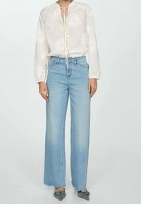 Bílá blůza se květinovou výšivkou, zapínáním vpředu na knoflíky a stahovacími šňůrkami u výstřihu, kombinovaná se světle modrými širokými džíny s roztrhaným lemem.