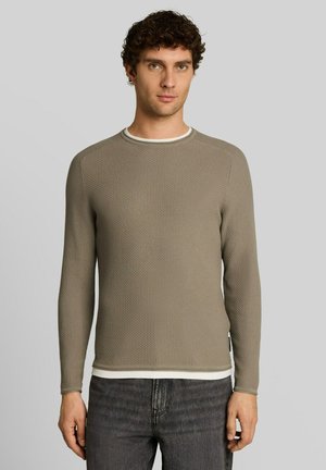 Jeune homme aux cheveux bouclés portant un pull kaki texturé à manches longues et un jean sombre, debout devant un fond gris clair uni.
