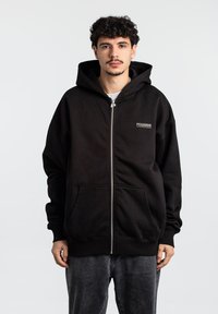 Sort zip-up hættetrøje lavet af bomuld, med en frontlomme og en stor hætte. "Pegador" logo i små bogstaver på den øverste venstre side.