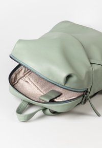 Grüner Kunstleder-Rucksack mit einem Reißverschluss, der ein gemustertes beiges Innenfutter und eine kleine Klappentasche im Inneren zeigt.