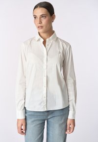 Camicia bianca con bottoni e maniche lunghe, realizzata in tessuto morbido, con un piccolo logo ricamato sul petto e colletto classico.
