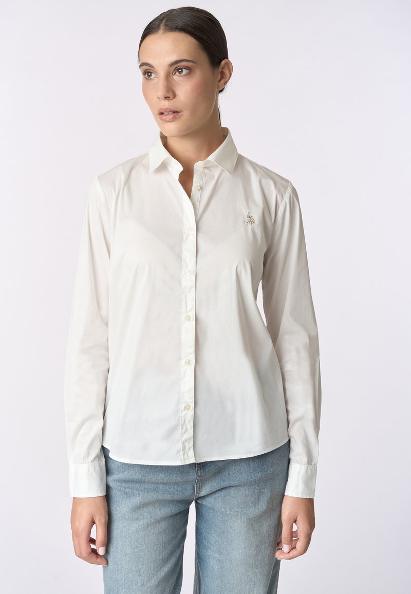 Camicia bianca con bottoni e maniche lunghe, realizzata in tessuto morbido, con un piccolo logo ricamato sul petto e colletto classico.