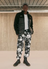 Schwarze, flauschige Jacke im Pufferdesign, graue Mütze, weißes T-Shirt, schwarz-weiß gemusterte Hose und schwarze Schnürstiefel.