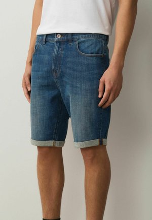 Man draagt middenblauwe spijkershorts met opgerolde zoom en voorzakken, gecombineerd met een wit ingestopt shirt.