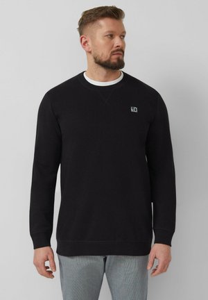 Mężczyzna o krótkich włosach i brodzie, ubrany w czarną koszulę z długim rękawem na białym podkoszulku oraz jasnoszare spodnie, patrzący w prawo.