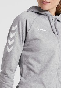 Haut à capuche zippé gris en matériau doux, avec une capuche, un logo blanc et des motifs en chevrons réfléchissants sur les manches.