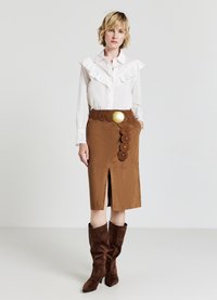 Gonna in suede marrone con spacco frontale e dettagli a cintura circolare; blusa bianca con della ruches sul davanti; stivali alti in suede marrone, dalla texture liscia.