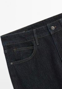 Donkerblauwe spijkerbroekband met voorkantzak, riemlussen, knoopsluiting en klinknageldetail op witte achtergrond.